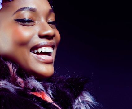 young pretty african american woman in spotted fur coat and flowers on head smiling on black background, fashion people concept close up