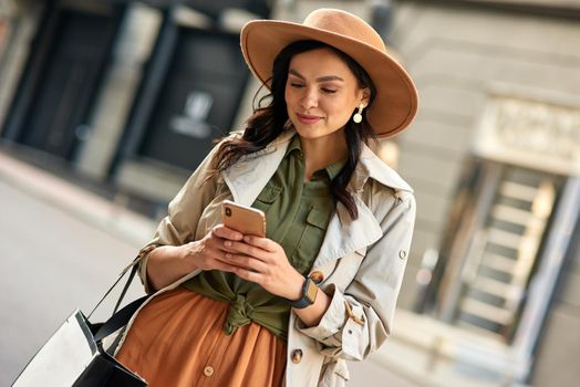 Chatting with friend. Young beautiful stylish woman wearing autumn coat and hat using her smartphone and smiling while standing on city street. Fashion, shopping, people lifestyle