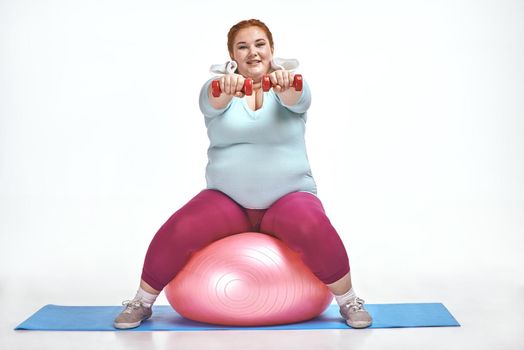 Funny picture of amusing, red haired, chubby woman on white background. Woman sitting on the ball and keep the dumbbells