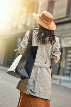 Rear view of a stylish woman wearing autumn grey coat and hat with big shopping bag walking city streets. Shopaholism, fashion, people lifestyle concept