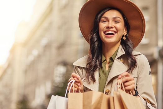 Shopping as therapy. Portrait of a young excited woman wearing grey coat and hat holding shopping bags, standing on city street and smiling at camera. Fashion, people lifestyle concept