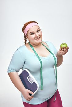 Funny picture of amusing, red haired, chubby woman on white background. Woman holding an apple and scales.