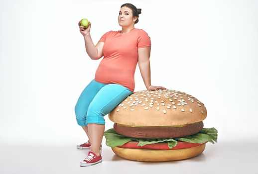 Funny picture of brunette, bearded, plump woman on white background. Woman wearing sportswear. She laid crouched on a huge sandwich