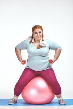 Funny picture of amusing, red haired, chubby woman on white background. Woman sitting on the ball and keep the dumbbells