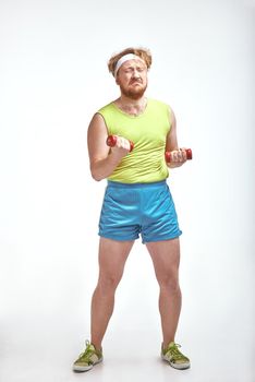 Funny picture of red haired, bearded, plump man on white background. Man wearing sportswear. Man holding the dumbbells