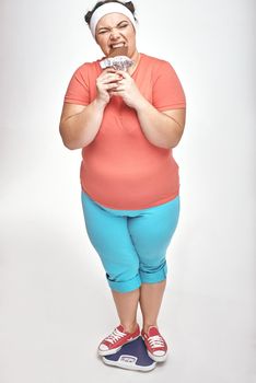 Funny picture of amusing, chubby woman on white background. She is eating chocolate while standing on the scales