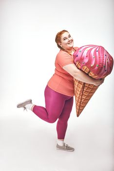 Funny picture of amusing, red haired, chubby woman on white background. Woman is holding a huge ice cream