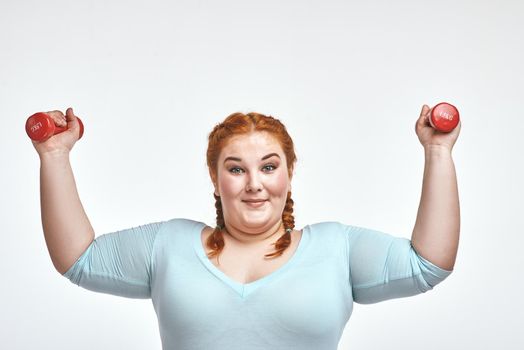 Funny picture of amusing, red haired, chubby woman on white background. Woman smiling and holding dumbbells