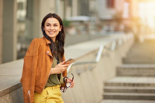 Side view of a young and happy cheerful business woman using her smartphone and smiling, looking aside while standing against blurred urban background outdoors, walking city streets. People lifestyle