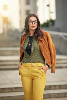 Vertical shot of a confident fashionable business woman wearing eyeglasses looking aside while standing against blurred urban background, walking city streets. People lifestyle concept