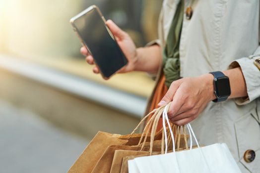 Cropped shot of a woman with shopping bags holding her smartphone while standing on city street outdoors. Shopaholism, sale, people lifestyle concept