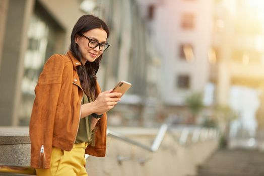 Side view of a young beautiful business woman using her smartphone, texting sms or chatting with friend while standing against blurred urban background outdoors, walking city streets. People lifestyle