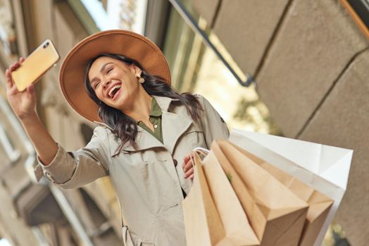 Young excited stylish woman with shopping bags standing near fashion boutique or shop and taking selfie in her smartphone, smiling at camera. Shopaholism, sale, people lifestyle concept