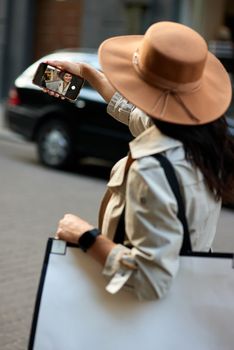 Rear view of a young stylish woman with big shopping bag making a selfie on her smartphone while walking city streets. Fashion, people lifestyle concept