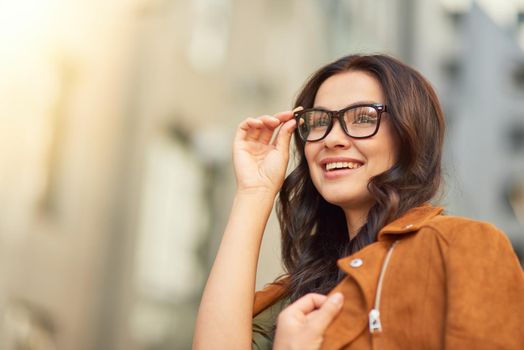 Portrait of a young cheerful business woman adjusting her eyeglasses and looking aside while standing against blurred urban background, walking city streets. People lifestyle concept