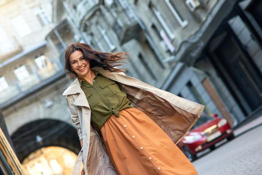 Young excited stylish caucasian woman wearing grey coat looking at camera and smiling while standing on the city street, walking outdoors on a warm autumn day. Beautiful people, fashion concept