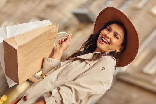 Positive emotions. Portrait of a young excited woman wearing grey coat and hat carrying shopping bags, standing on city street and smiling at camera. Fashion, people lifestyle concept