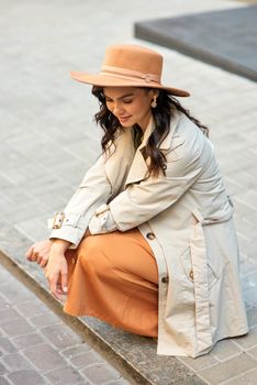 Vertical shot of a young attractive stylish woman wearing autumn coat and hat looking aside and smiling while squatting down on the city street. People lifestyle concept