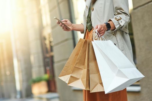 Texting sms to a friend. Cropped shot of a woman with shopping bags using her smartphone while standing on city street outdoors. Shopaholism, sale, people lifestyle concept