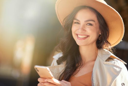 Chatting with friend. Portrait of a young beautiful and happy caucasian woman wearing hat using smartphone and smiling at camera while standing on the city street on a sunny day