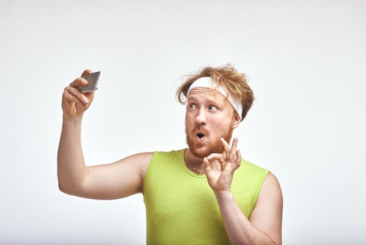 Funny picture of red haired, bearded, plump man on white background. Man wearing sportswear. Man smiling and taking a selfie