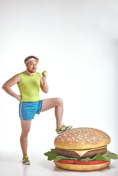 Funny picture of red haired, bearded, plump man on white background. Man wearing sportswear. Man put his leg to a huge sandwich