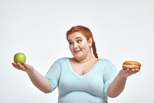 Funny picture of amusing, red haired, chubby woman on white background. Woman holding apple and sandwich. She is looking at an apple