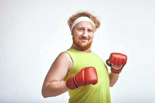 Funny picture of red haired, bearded, plump man on white background. Man wearing sportswear and red boxing gloves