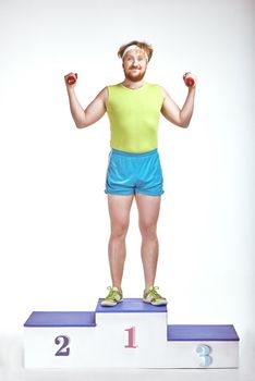 Funny picture of red haired, bearded, plump man on white background. Man wearing sportswear. He is standing on a pedestal