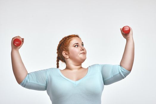 Funny picture of amusing, red haired, chubby woman on white background. Woman smiling and holding dumbbells