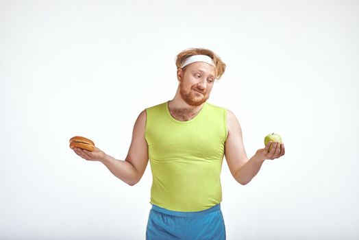 Funny picture of red haired, bearded, plump man on white background. Man wearing sportswear. Man holding apple and sandwich