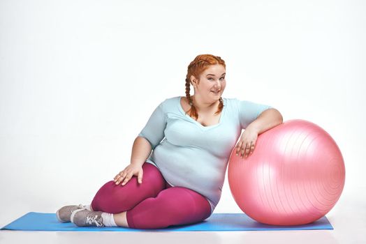 Funny picture of amusing, red haired, chubby woman on white background. Woman trains on the mat, she leans on the ball