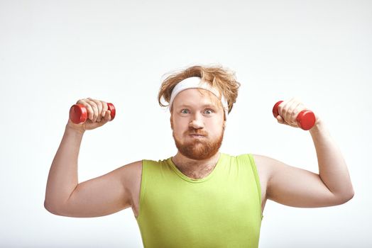 Funny picture of red haired, bearded, plump man on white background. Man wearing sportswear. Man holding the dumbbells