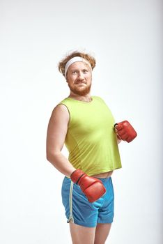 Funny picture of red haired, bearded, plump man on white background. Man wearing sportswear and red boxing gloves