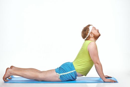 Funny picture of amusing, red haired, chubby man on white background. Man trains on the mat.