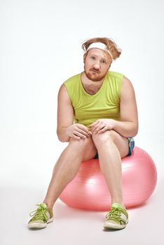 Funny picture of red haired, bearded, plump man on white background. Man wearing sportswear. Man sitting on a ball.