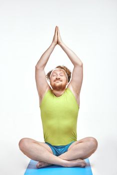 Funny picture of red haired, bearded, plump man on white background. Man wearing sportswear. Man sitting on the mat, he is meditating