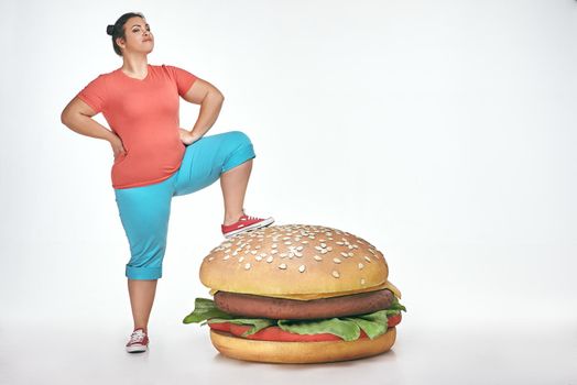 Funny picture of brunette, bearded, plump woman on white background. Woman wearing sportswear. She put her leg to a huge sandwich