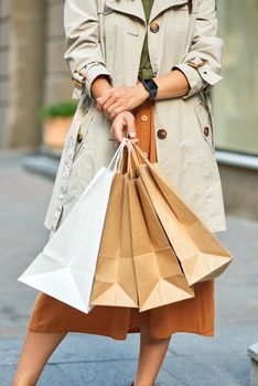Cropped shot of a woman wearing grey autumn coat with shopping bags standing on city street outdoors. Shopaholic, fashion, people lifestyle concept
