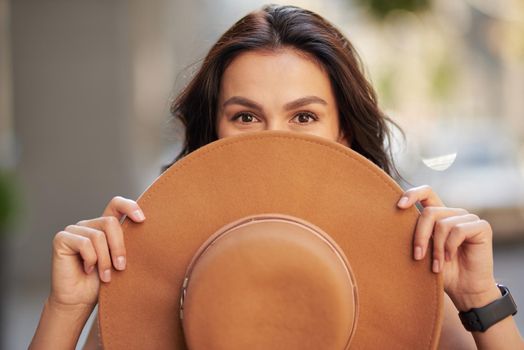 Portrait of a young playful caucasian woman covering her face with hat, looking at camera while standing outdoors. People lifestyle and fashion concept