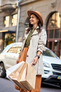 Young happy beautiful and stylish caucasian woman shopping bags wearing autumn grey coat and hat looking aside and smiling while standing on the city street. Shopaholism, fashion, people lifestyle