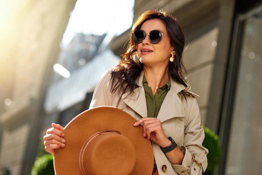 Young stylish woman wearing eyeglasses holding hat and looking aside while walking city streets on a warm autumn day. Fashion, people lifestyle concept