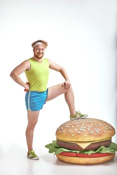 Funny picture of red haired, bearded, plump man on white background. Man wearing sportswear. Man put his leg to a huge sandwich
