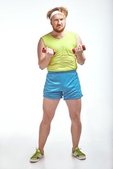 Funny picture of red haired, bearded, plump man on white background. Man wearing sportswear. Man holding the dumbbells