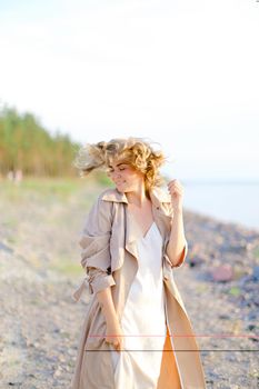 Young smiling girl standing on sea beach and wearing coat with white dress. Concept of enjoying freedom and summer photo session.