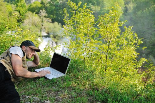 young businessman with hat work on thin laptop outdoor in nature with beautiful waterfalls in background representing freedom and wireless technology concept 