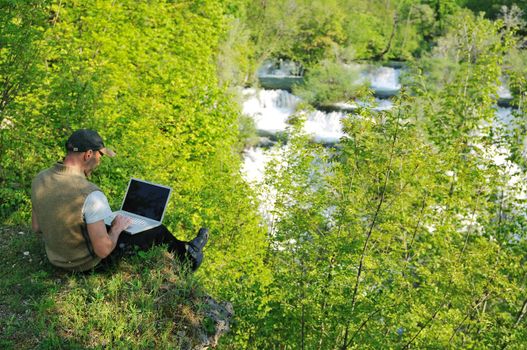 young businessman with hat work on thin laptop outdoor in nature with beautiful waterfalls in background representing freedom and wireless technology concept 