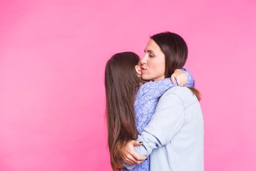 people, happiness, love, family and motherhood concept - happy little daughter hugging and kissing her mother over pink background.