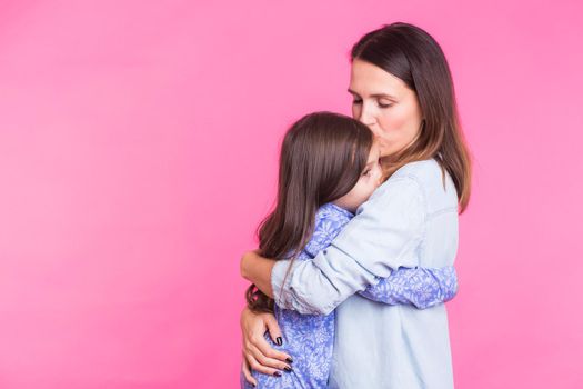 people, happiness, love, family and motherhood concept - happy little daughter hugging and kissing her mother over pink background.