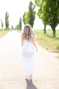 Back view of blonde bride wearing white dress walking on road. Concept of wedding photo session and fiancee.